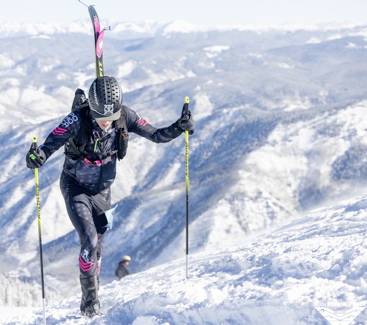 Solo racer bootpacking steep slope with poles and mountain range behind