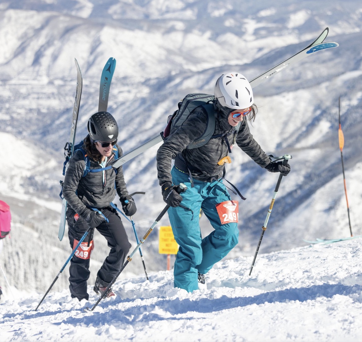 Two skimo racers bootpacking uphill with skis on packs during race day